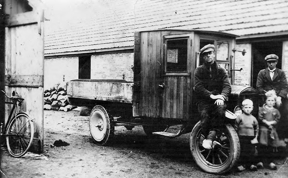 Historische zwart-witfoto van een vrachtwagen met houten cabine voor een schuur